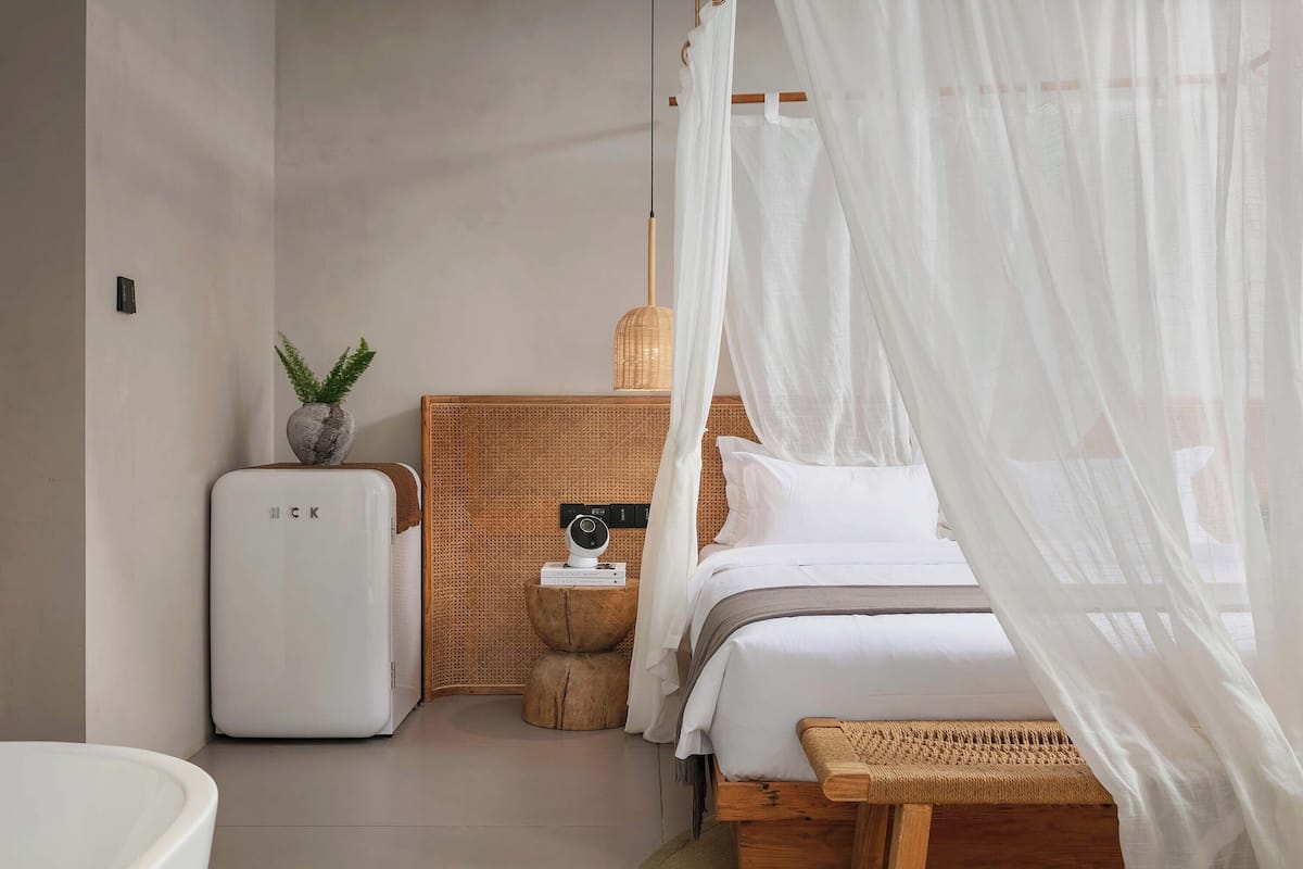 Un dormitorio con cama, mesita de noche, un pequeño calefactor blanco, lámpara de pared, cesta tejida y una planta.