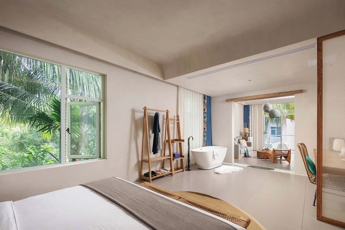 Un dormitorio moderno con un ventanal grande, una cama, un perchero de madera con ropa, una bañera y vistas al exterior.