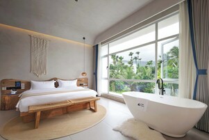 Un dormitorio moderno con una cama grande, un banco de madera, una bañera blanca y vistas a palmeras a través de amplios ventanales.