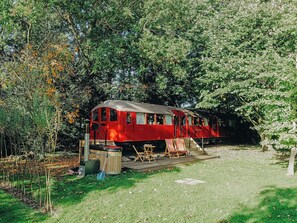 Um vagão de trem vermelho com cobertura, localizado em área arborizada com grama e árvores ao redor.