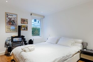 Un dormitorio con cama, mesita de noche, lámpara y ventana con vista a un edificio.