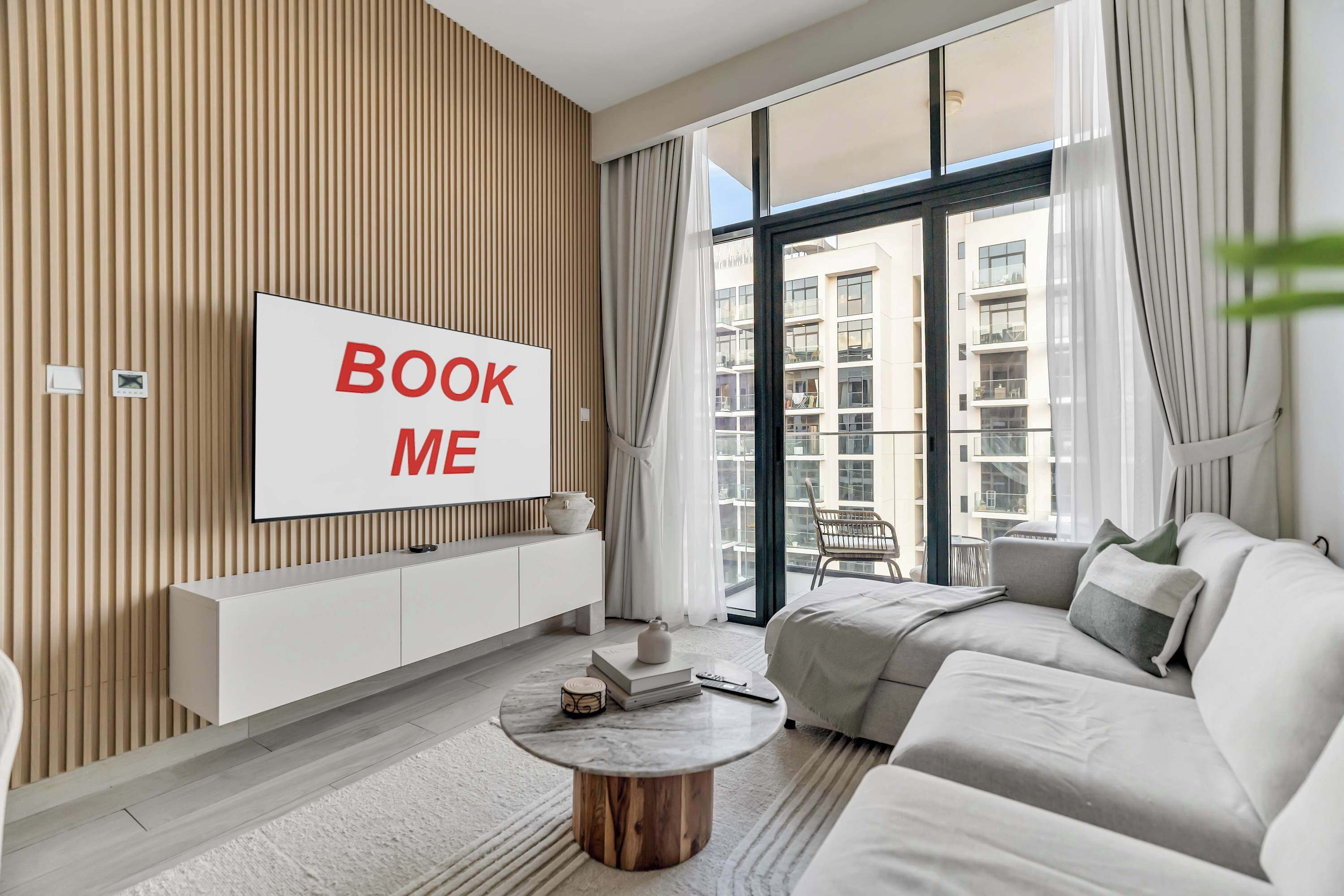 A modern living room with a large TV displaying 'BOOK ME,' a sofa, a coffee table, and a view of a building through large windows.