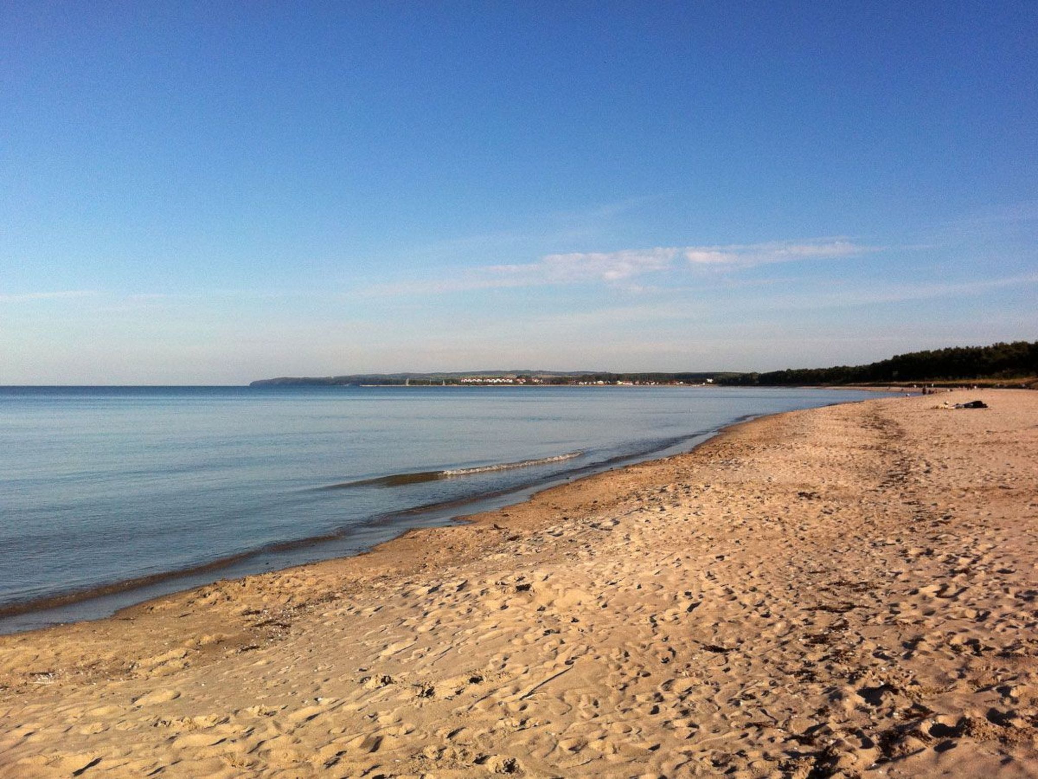 Ampia spiaggia sabbiosa con onde calme, cielo sereno e una costa lontana con edifici.