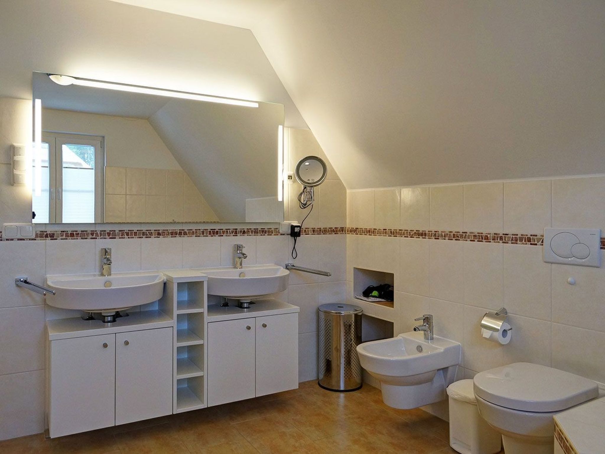 A modern bathroom with two sinks, a bidet, and a toilet. There is a mirror above the sinks, and a trash can is visible.