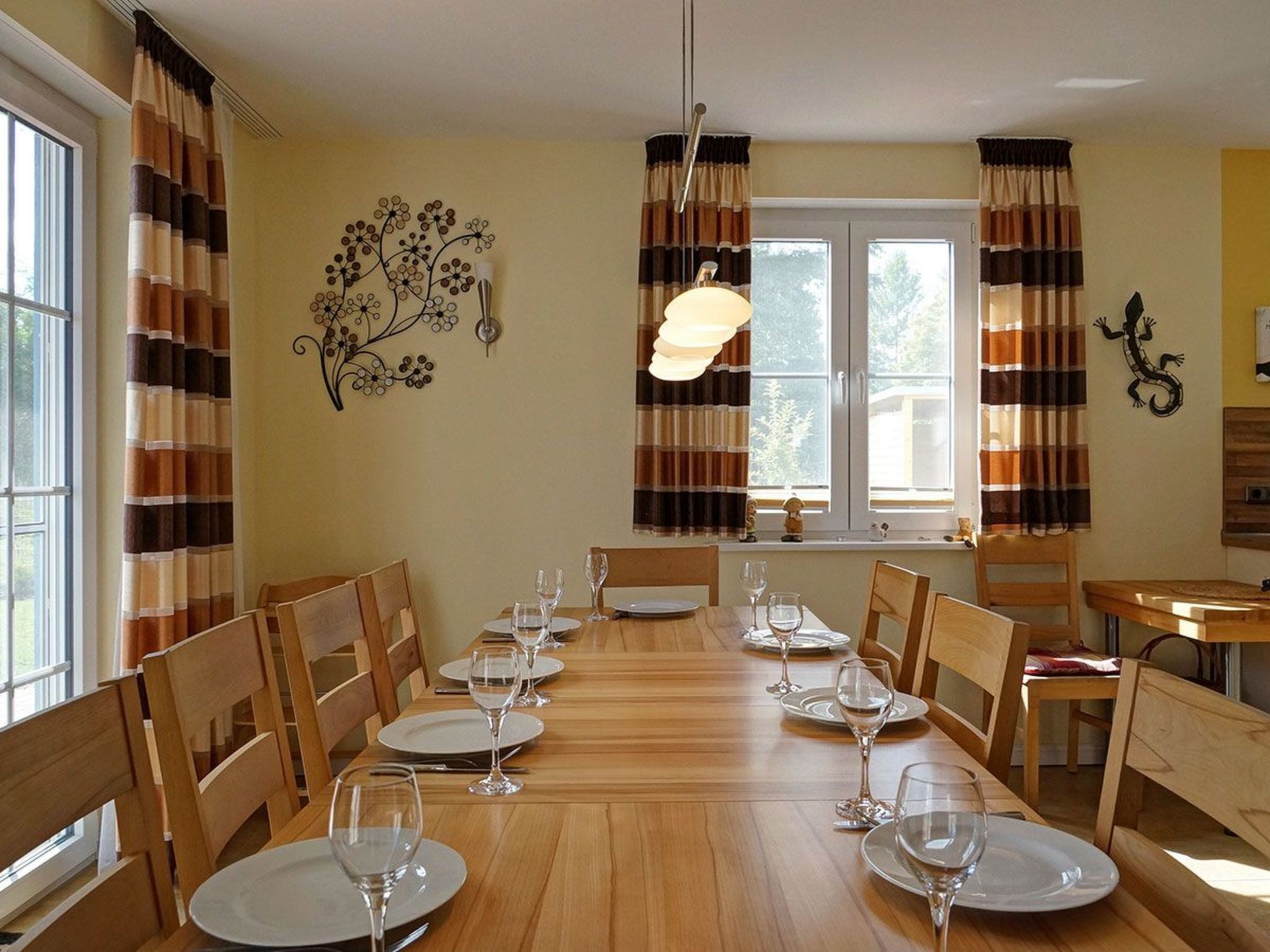 Comedor con una larga mesa de madera dispuesta para seis personas, rodeada de sillas de madera. Hay dos ventanas con cortinas, una lámpara fijada a la pared y un adorno en la pared.