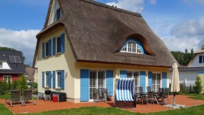 Una casa con techo de paja, contraventanas azules y un pequeño patio con mobiliario exterior.