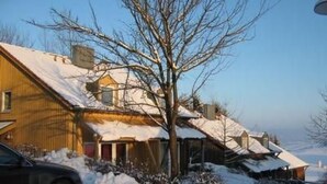Ein Wohnhaus mit geneigtem, mit Schnee bedecktem Dach, einem sichtbaren Schornstein und einem geparkten Auto davor.