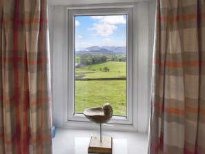 A window with a view of a green field and mountains, curtains, and a sculpture on a windowsill.
