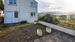 Une maison à deux étages, à l’extérieur blanc, avec un petit jardin devant, agrémenté de pas japonais, et offrant une vue sur des collines lointaines.