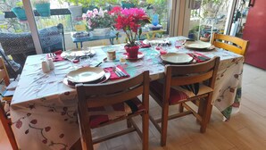 Une table à manger dressée avec des assiettes, des couverts et des verres, entourée de chaises, agrémentée de plantes en pot et offrant une vue sur la verdure à l’extérieur.
