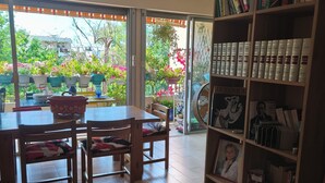 Un coin repas avec une table et des chaises en bois, une bibliothèque remplie de livres et un tableau encadré, et une vue sur un jardin à travers des portes vitrées.