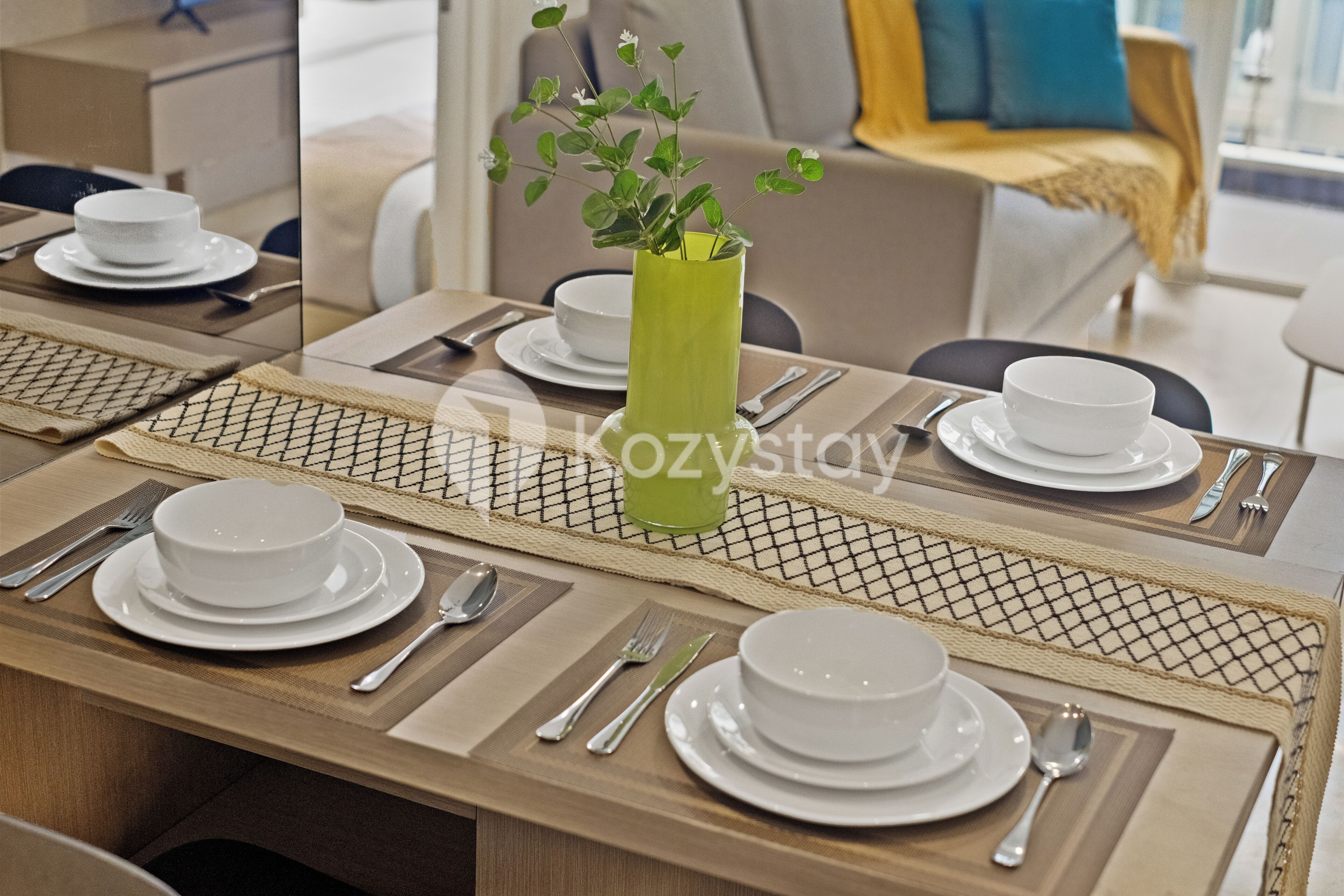 A dining table set with white plates, bowls, and cutlery, a green vase with a plant, and a woven placemat.