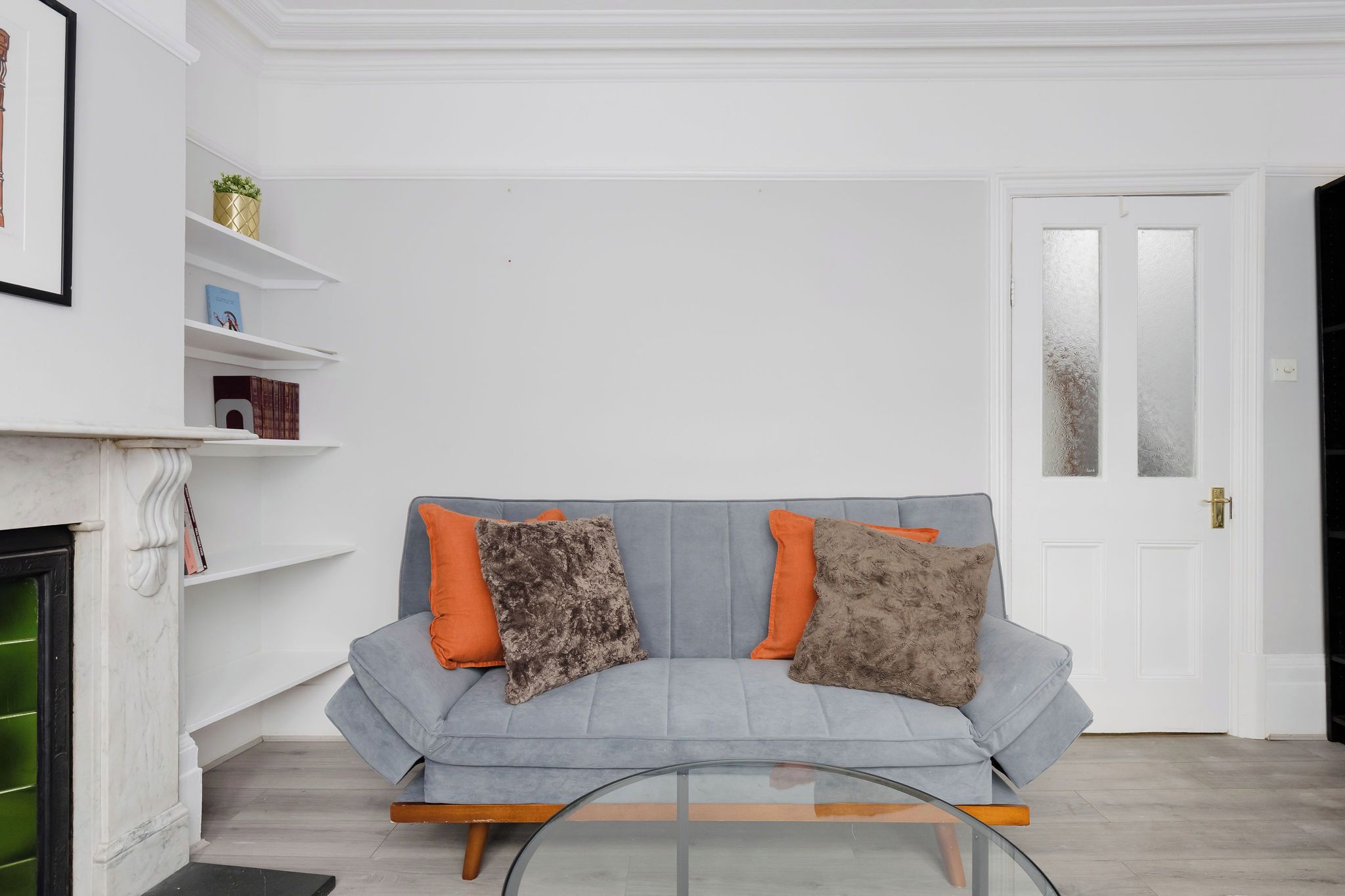 Een woonkamer met een grijze bank, twee oranje en twee grijze kussens, een glazen salontafel, een witte haard en een witte deur met geëtste glaspanelen.