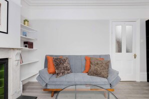 Een woonkamer met een grijze bank, oranje kussens, een open haard en een witte deur.