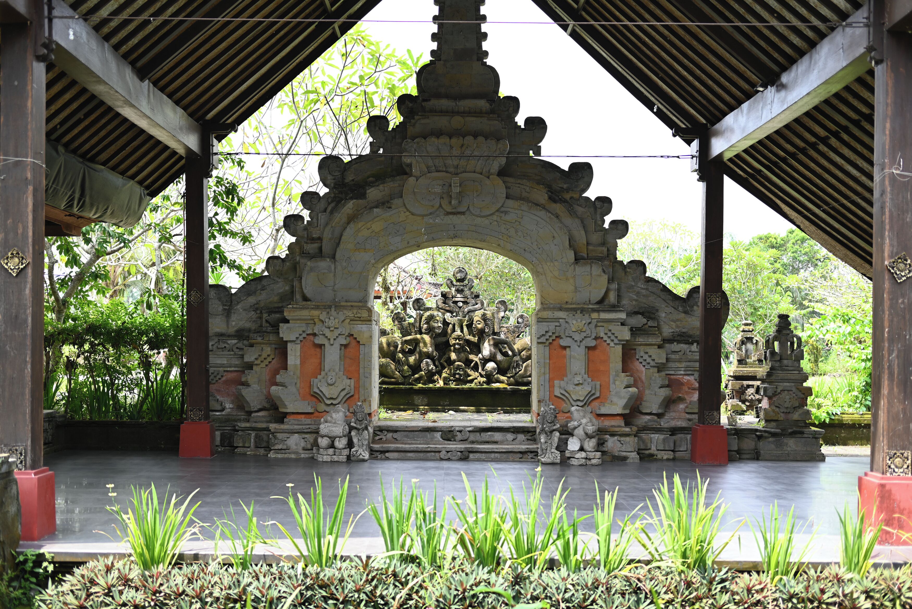 A stone structure with an archway, surrounded by greenery and statues.