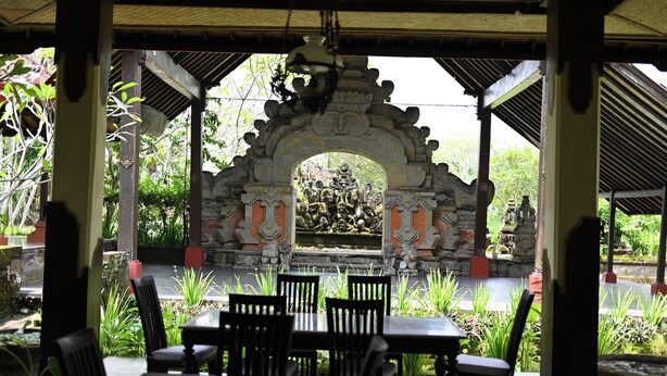 A covered outdoor seating area with stone arches, tables, and chairs, surrounded by lush greenery. - Suly Resort Ubud (Ubud)