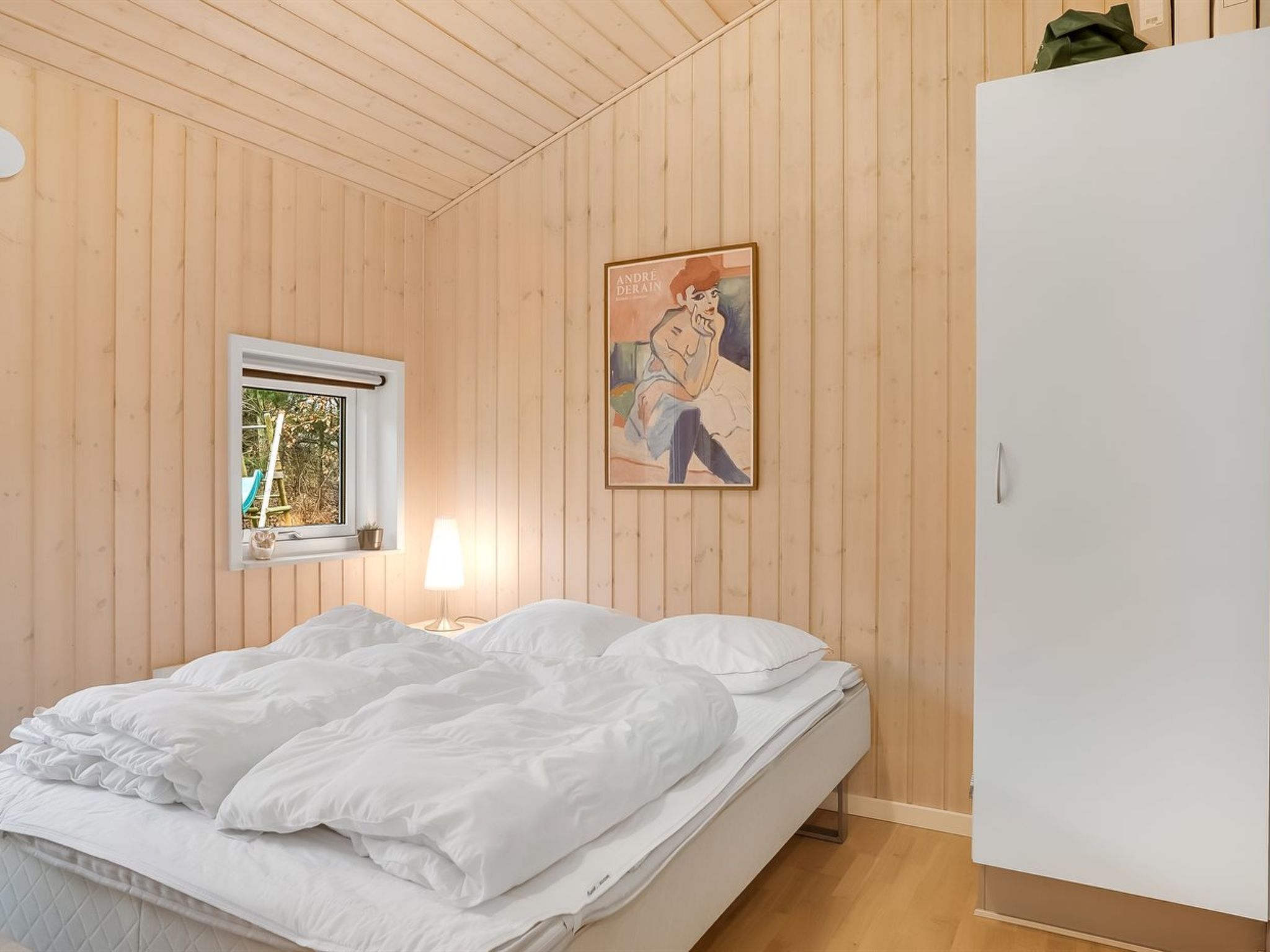A bedroom with a bed, a window, a lamp, and a wall with a framed picture.