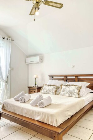 Un lit bien fait, avec du linge de lit et des serviettes blanches, une tête de lit en bois, une table de chevet avec une lampe et un appareil de climatisation fixé au mur.