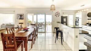 A spacious dining area with a wooden table and chairs, a kitchen with modern appliances, and a view of the sea through large sliding doors. - Villa Bel Ocean - Piscine & Vue Mer Saint Gilles (Saint-Paul)