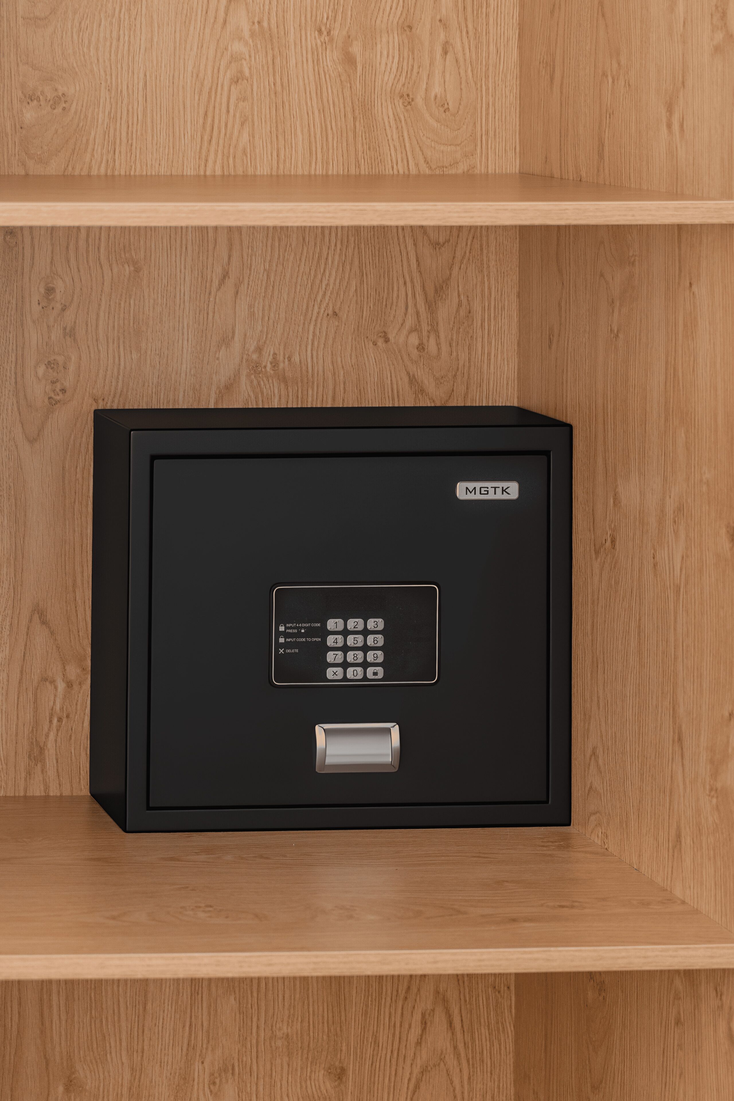 A black safe with a keypad and a brand label 'MGTK' on a wooden shelf.