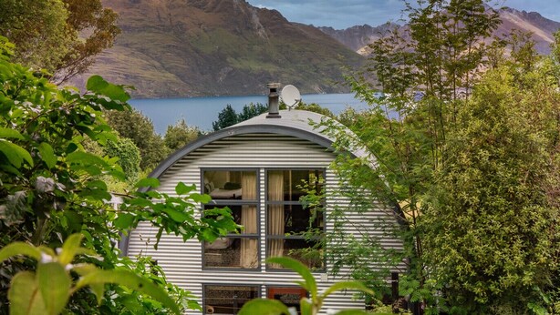 A small, single-story cabin with a curved roof, surrounded by lush greenery and overlooking a lake with mountains in the background. - Arc House | 5min to Town • Spacious Loft House (Queenstown)