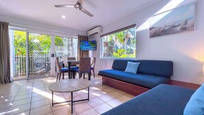 Ein modernes Wohnzimmer mit einer Couch, einem Couchtisch und einem Fernseher. Ein Balkon bietet Blick auf Grünflächen.