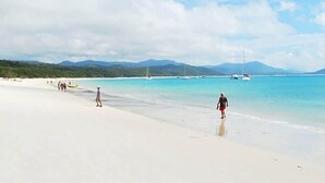 Ein Strand mit klarem türkisblauem Wasser, ein paar Spaziergänger und Segelboote in der Ferne.