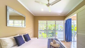 A hotel room with a bed, a ceiling fan, a large window with blinds, and a view of greenery outside. - Poolside Garden Apt at Mango House, Whitsundays (Airlie Beach)