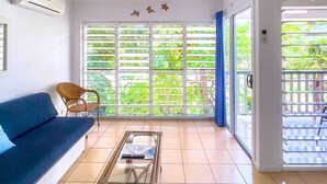 A living room with a blue sofa, a glass-top coffee table, a ceiling fan, and a view through large windows to a balcony.