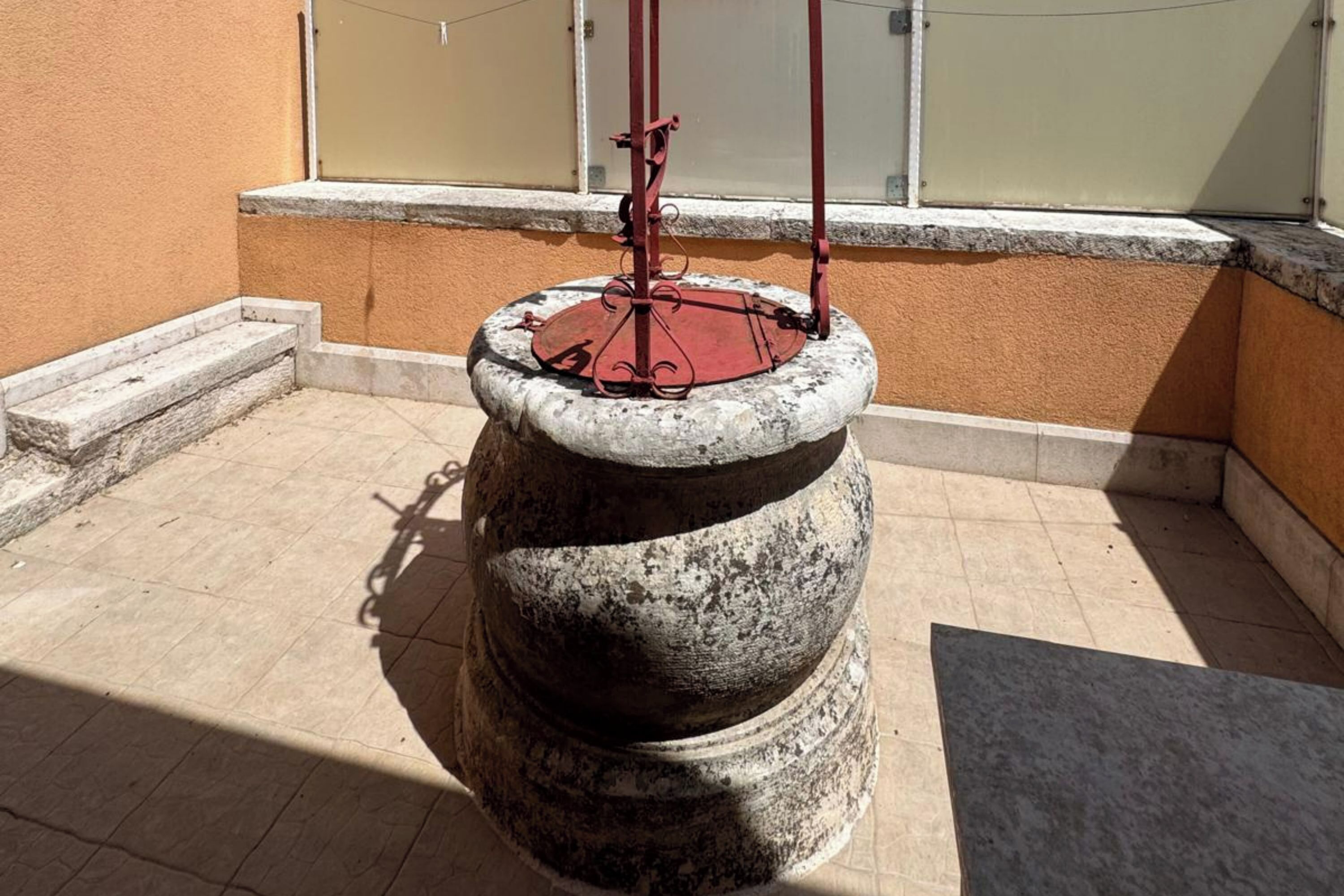 En stenvandpost med metalstænger og en rød sokkel, omgivet af en fliseterrasse og en beige mur.