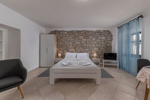 A bedroom with a stone wall, a bed with white linens, a TV on a stand, and a chair. - Iva Apartments (Bale)