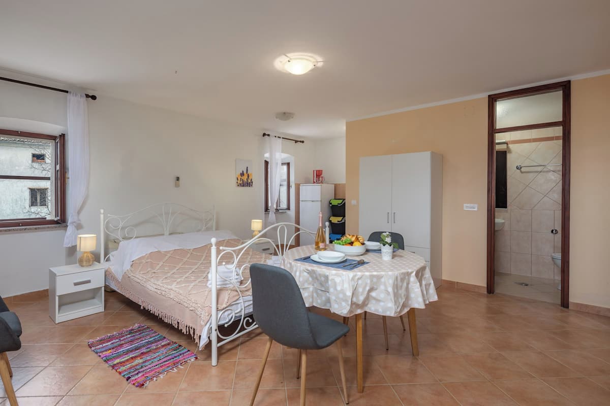 Une chambre à coucher avec un lit, une table de chevet et une petite table avec une chaise. Il y a une fenêtre avec des rideaux, une porte donnant sur une salle de bain et un sol carrelé.