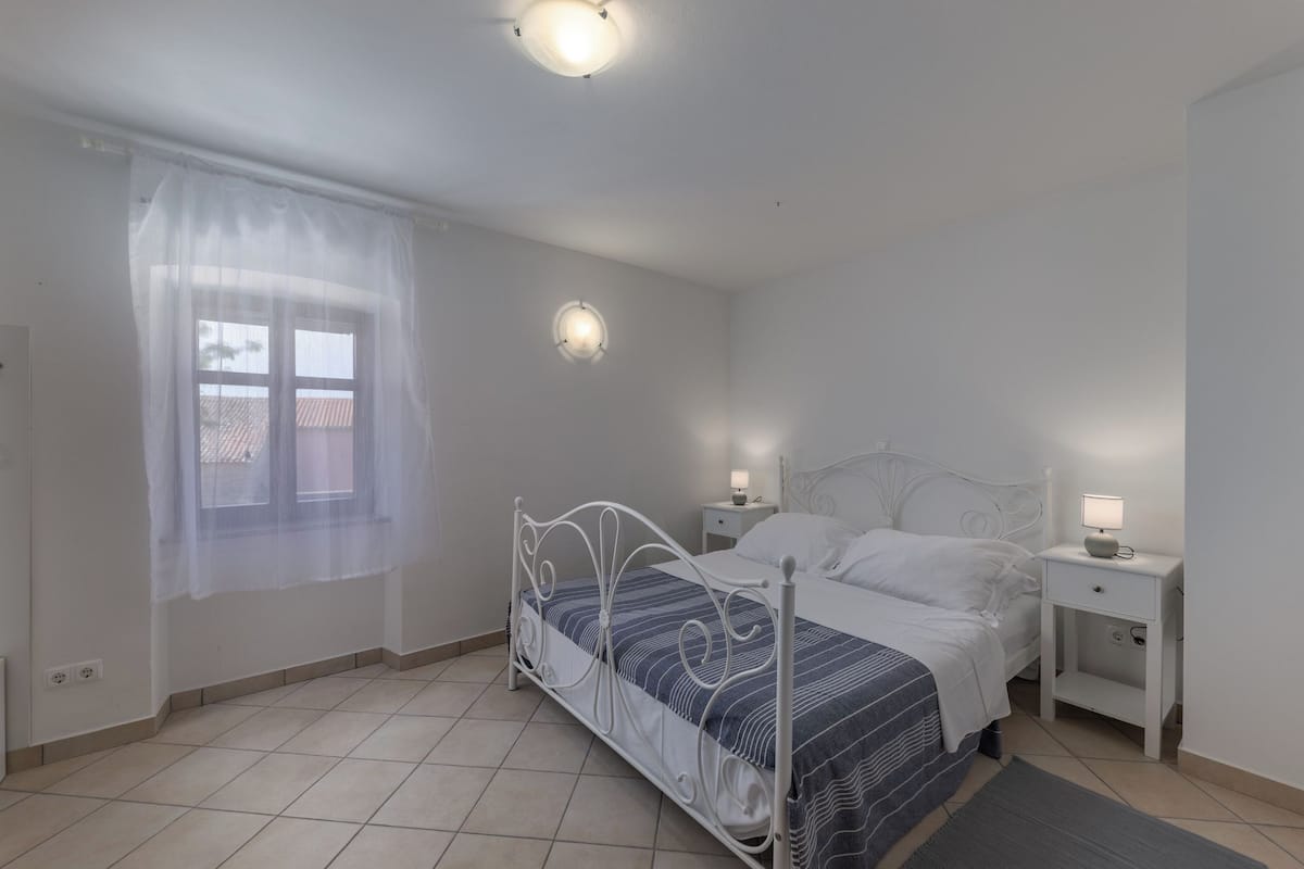 Une chambre à coucher comprenant un lit, des tables de chevet, une fenêtre avec un rideau léger et un luminaire fixé au mur.