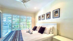 Une chambre à coucher comprenant un lit, une table de chevet, un ventilateur de plafond et une fenêtre avec des stores.