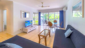 A modern living room with a flat-screen TV, a sofa, a coffee table, and a dining area with chairs. - Poolside Whitsundays apartment at Mango House (Airlie Beach)