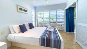 A bedroom with a bed, pillows, a ceiling fan, a window with blinds, and a view of greenery outside. - Poolside Mango House Apartment, Whitsundays (Airlie Beach)