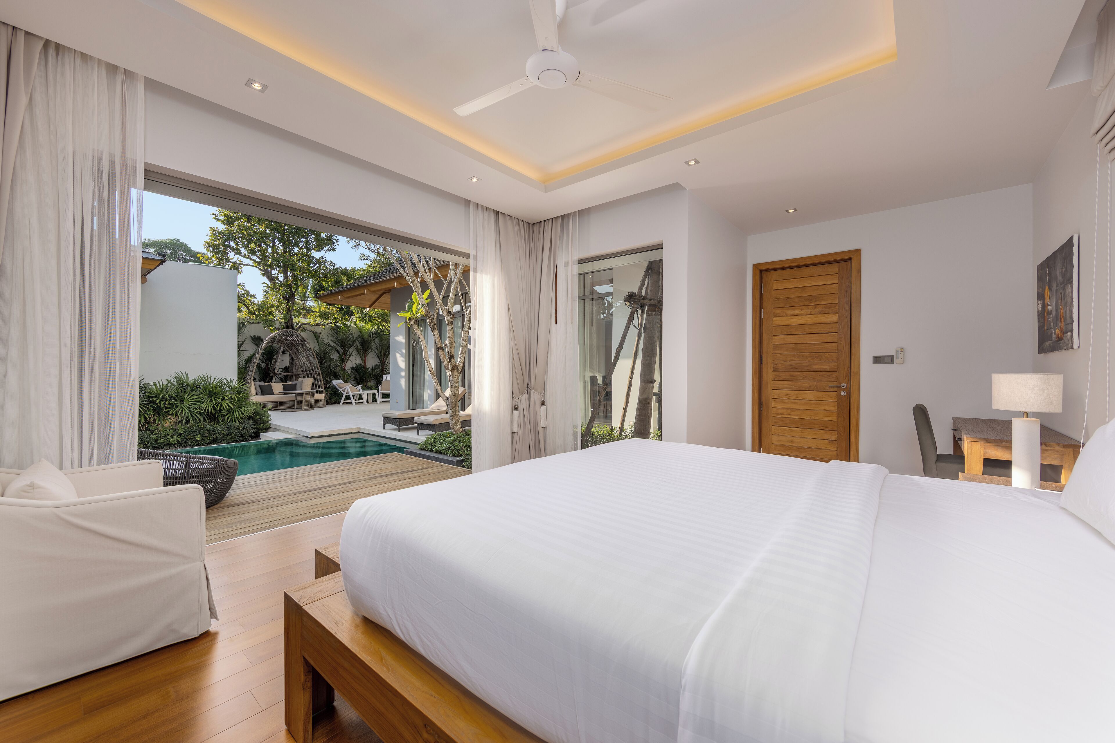 Une chambre dotée d’un grand lit, d’un banc en bois, d’un bureau avec une lampe, et offrant une vue sur une piscine et un espace extérieur.