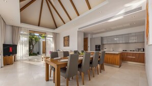 A modern kitchen with wooden cabinets and a central island, a dining area with a wooden table and grey chairs, and a TV on the left. - Modern 3BR Villa Kaimana Private Pool (Thep Krasattri)