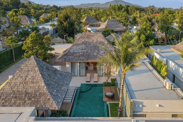 A resort with multiple bungalow-style buildings, each with a thatched roof and surrounded by lush greenery. A swimming pool is centrally located, with outdoor seating and a palm tree nearby. - Modern 3BR Villa Kaimana Private Pool (Thep Krasattri)