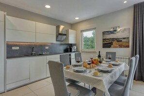 A modern kitchen with integrated appliances, a dining area with a table set for four, and a view of the outdoors through a window. - Elegant Panoramic Seaview Apartments (Medulin)