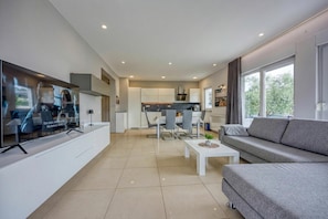 Een moderne woonkamer met een flatscreen televisie, een grijze bank, een salontafel, en een eethoek met een witte tafel en stoelen.