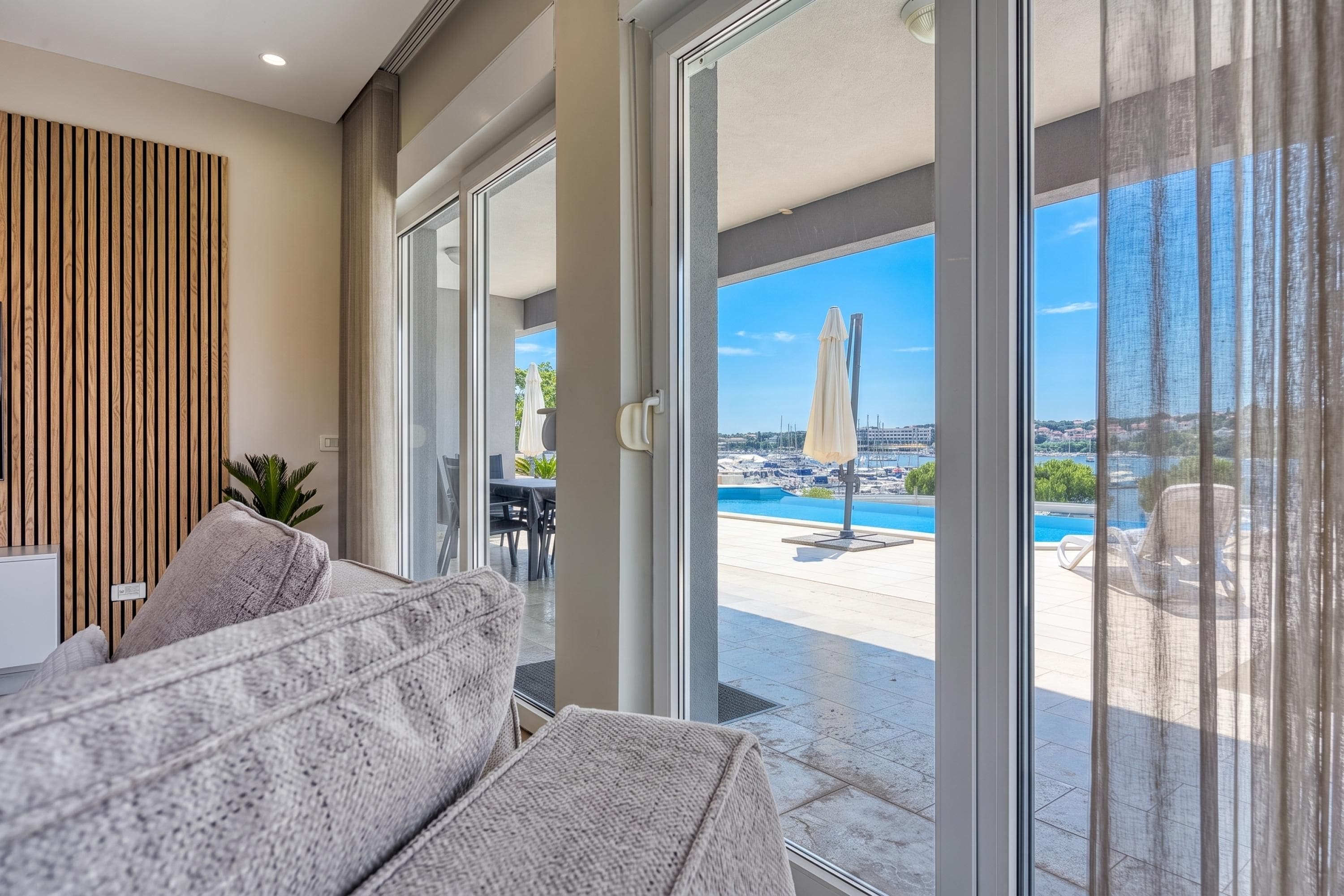 Un salón moderno con sofá, pared de paneles de madera y vista a una piscina exterior y un puerto deportivo.