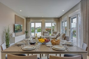 A dining area with a table set for six, a fruit bowl, and a bottle of wine. - Elegant Panoramic Seaview Apartments (Medulin)