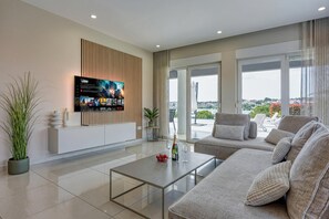A modern living room with a flat-screen TV, a sofa, a coffee table, and a view of the city through large windows. - Elegant Panoramic Seaview Apartments (Medulin)