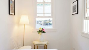 Quarto com cama, abajur, mesinha com um vaso de flores e janela com vista para uma cidade.