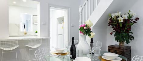 A modern dining area with a glass table set for four, white chairs, and a white staircase in the background.