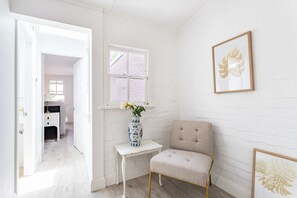 Une pièce avec un mur en briques blanches, une fenêtre donnant sur un beau paysage, une chaise, une petite table avec un vase et un tableau encadré.