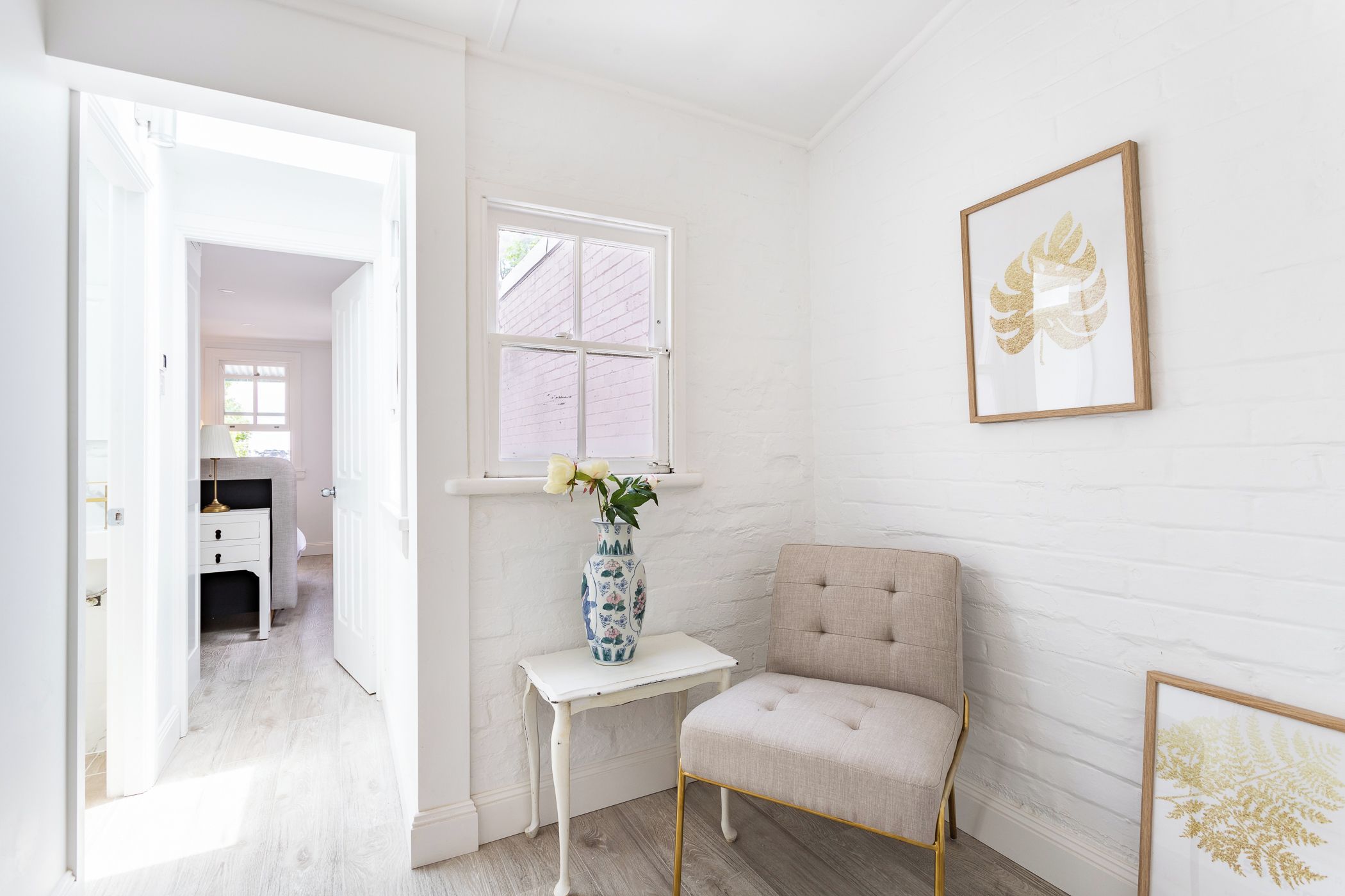 A room with a white brick wall, a window with a view, a chair, a small table with a vase, and a framed picture.