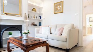 Uma sala de estar com sofá branco, mesa de centro de madeira, lareira e estante com objetos decorativos.