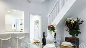 Uma área de jantar moderna com mesa de vidro, cadeiras de arame brancas e um vaso branco com flores.